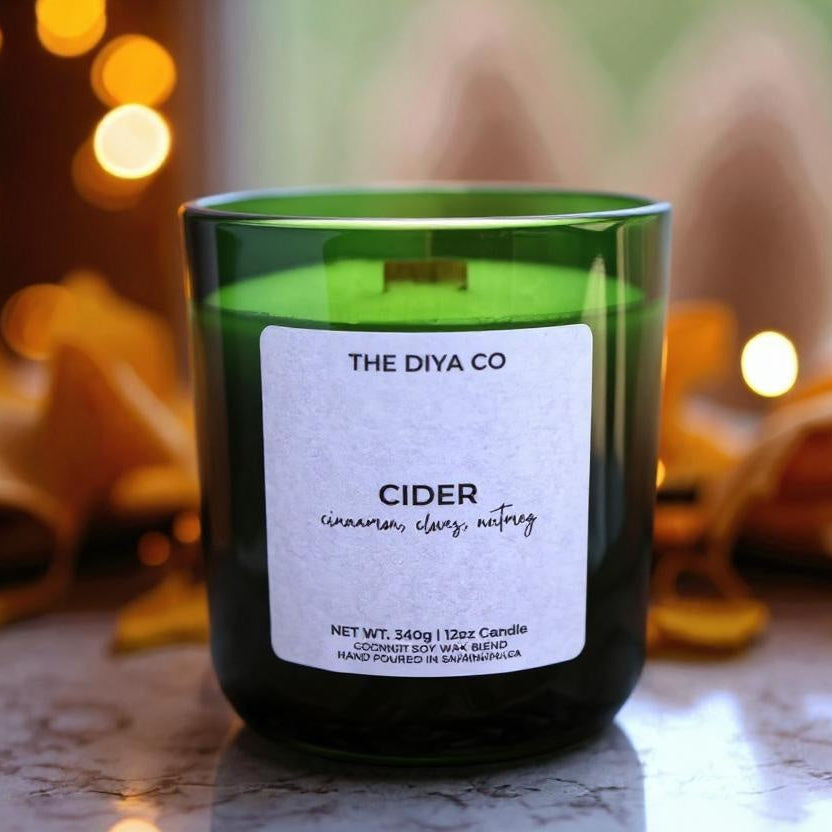 Green candle labeled 'The Diya Co Cider' with blurred lights and leaves in the background