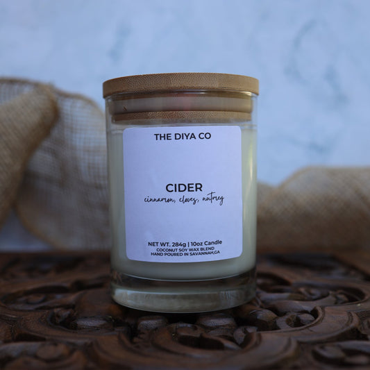 Candle jar labeled 'The Diya Co Cider' on a decorative wooden surface with burlap in the background.