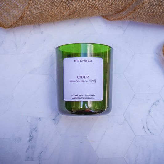 Green candle labeled 'Cider' from 'The Dya Co.' on a marble surface with burlap fabric.