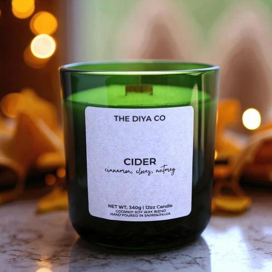 Green candle labeled 'The Diya Co Cider' with blurred lights and leaves in the background