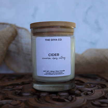 Candle jar labeled 'The Diya Co Cider' on a decorative wooden surface with burlap in the background.