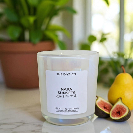 Candle labeled 'Napa Sunsets' by The DIYA Co. on a surface with figs and a pear, and a plant in the background.
